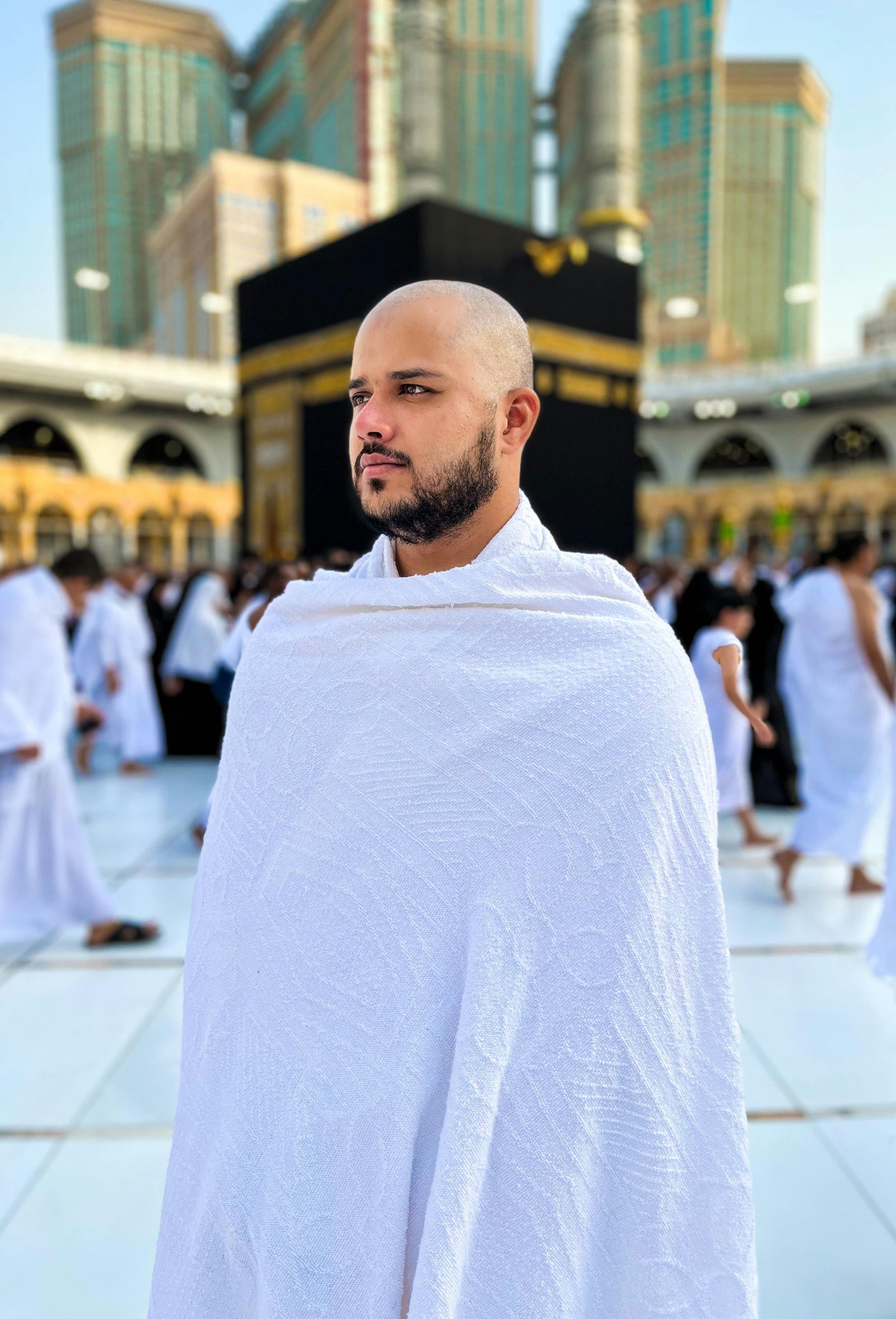 A man with a shaved head and beard, wearing white ihram clothing, standing at the Kaaba during Hajj pilgrimage in Mecca, Saudi Arabia, with a background of other pilgrims and high-rise buildings.