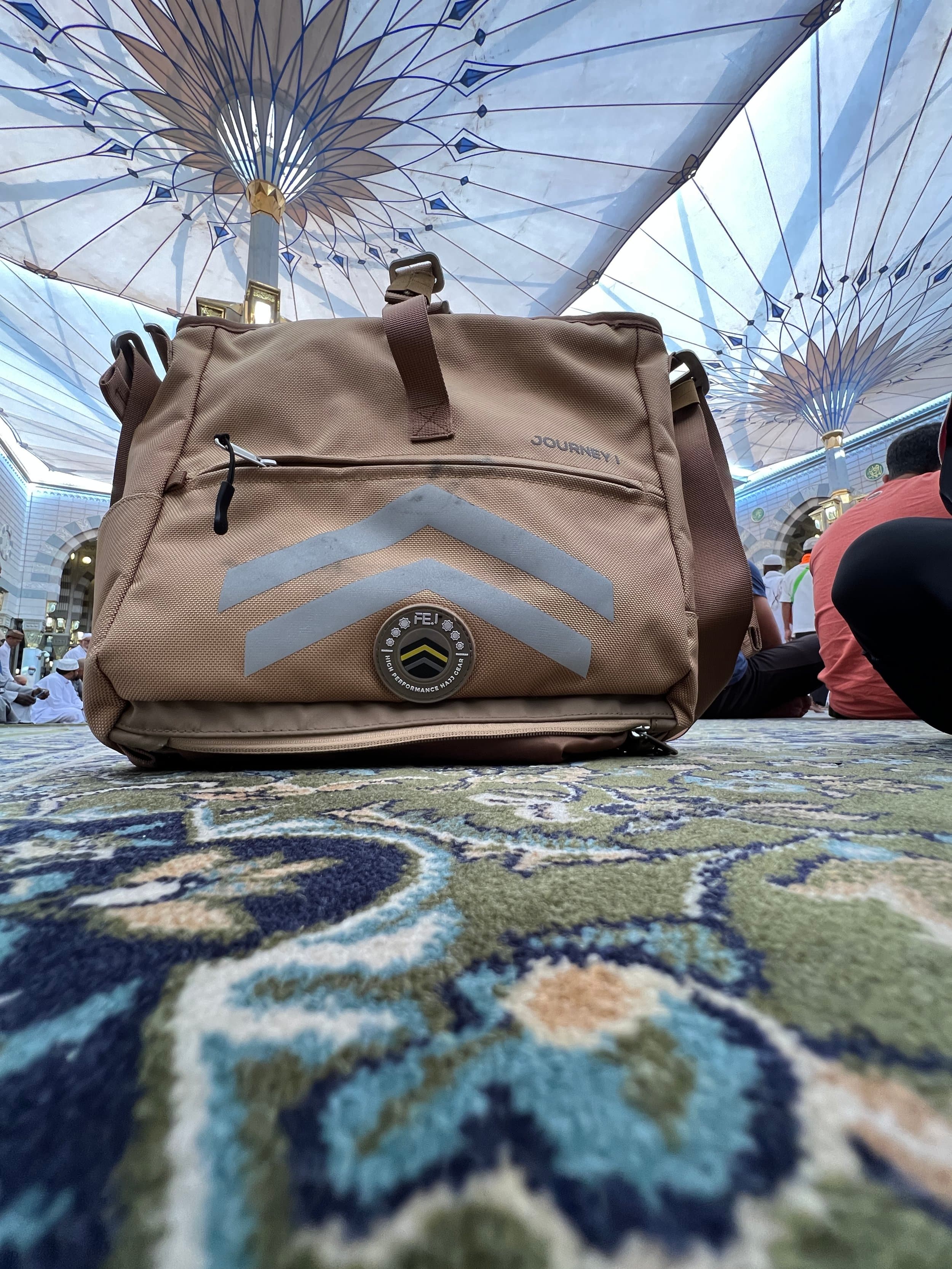 Close-up of a beige travel bag with reflective chevron markings and a round badge, placed on a patterned carpet in an ornate mosque, with mosque's decorative ceiling and people in the background.