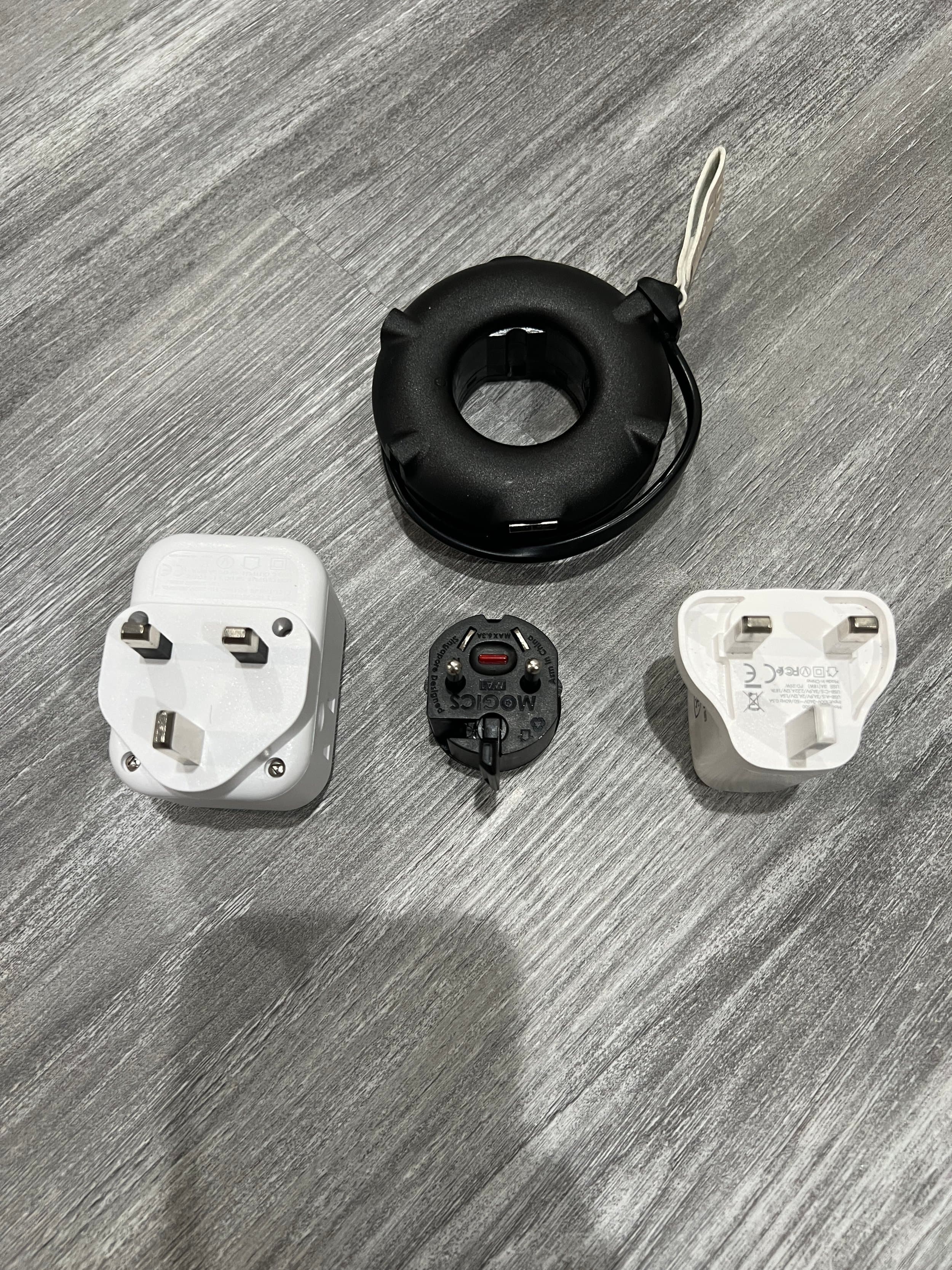 Electrical components including a white power plug, a black plug adapter, a white socket, and a black circular device on a gray wooden surface.