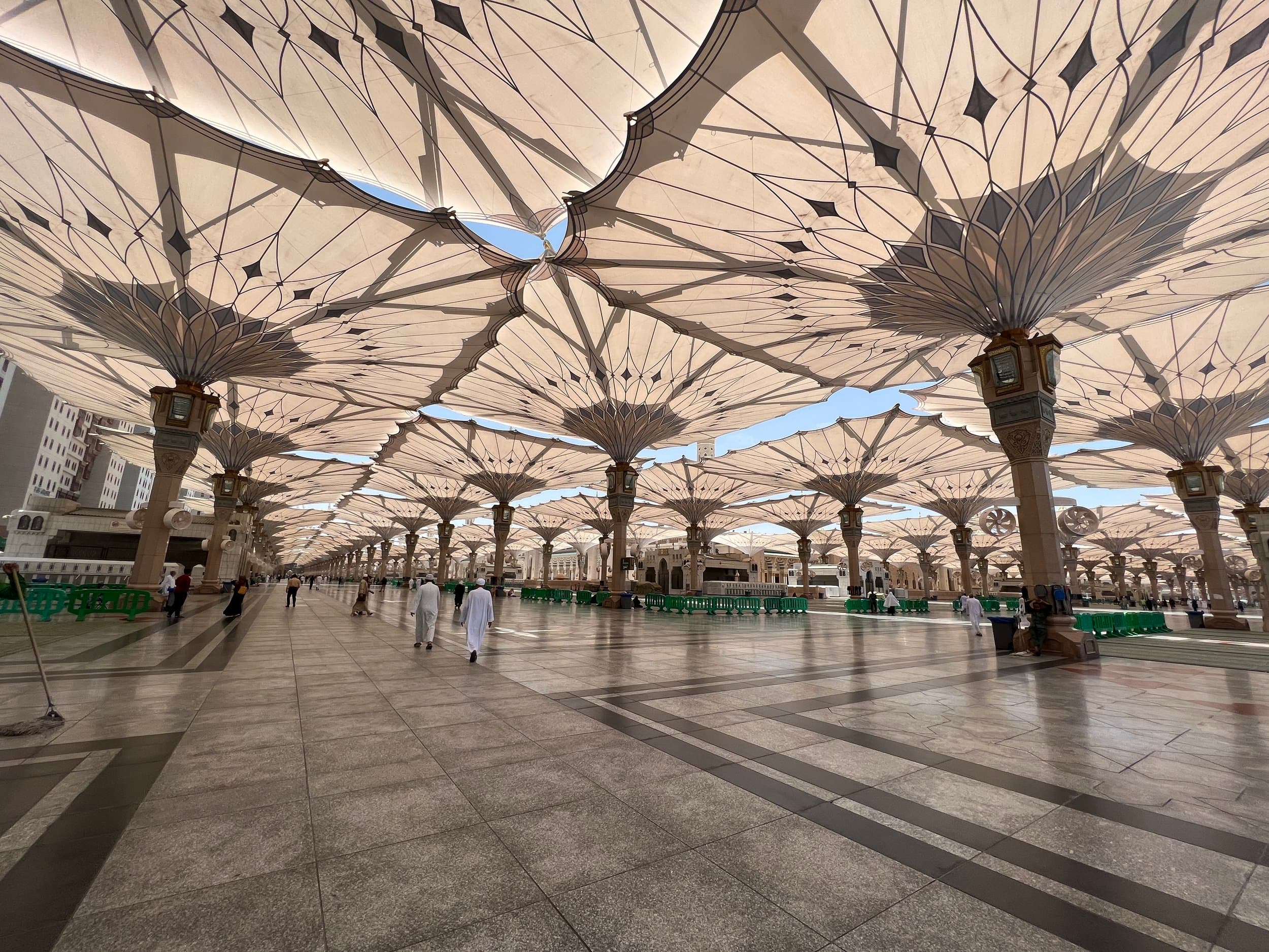 Large open mosque courtyard with multiple beige umbrellas overhead, supported by decorated columns, and several people walking across the marble floor.