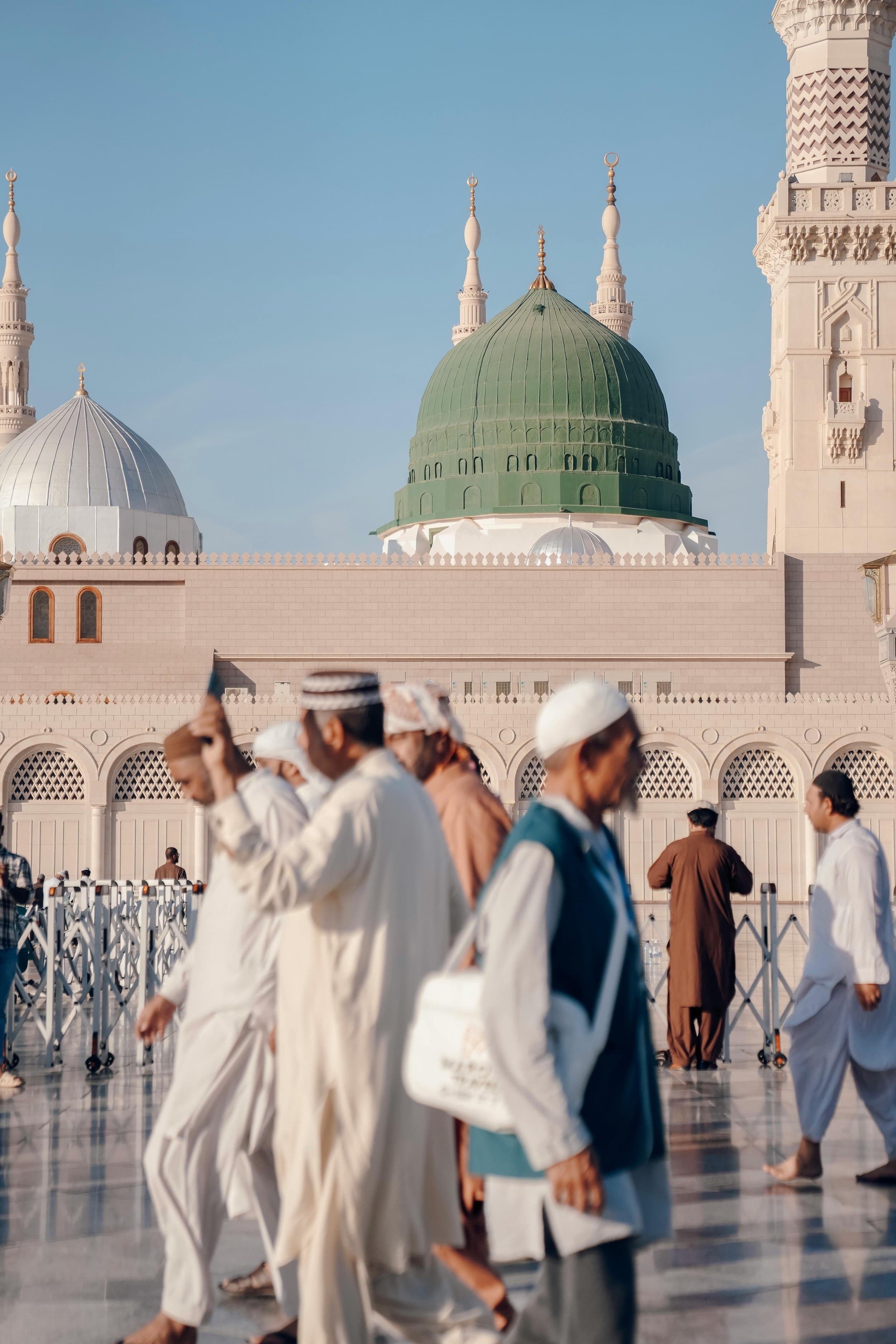 People walking outside the Al-Masjid an-Nabawi mosque with green dome and minarets in the background, in Medina, Saudi Arabia.