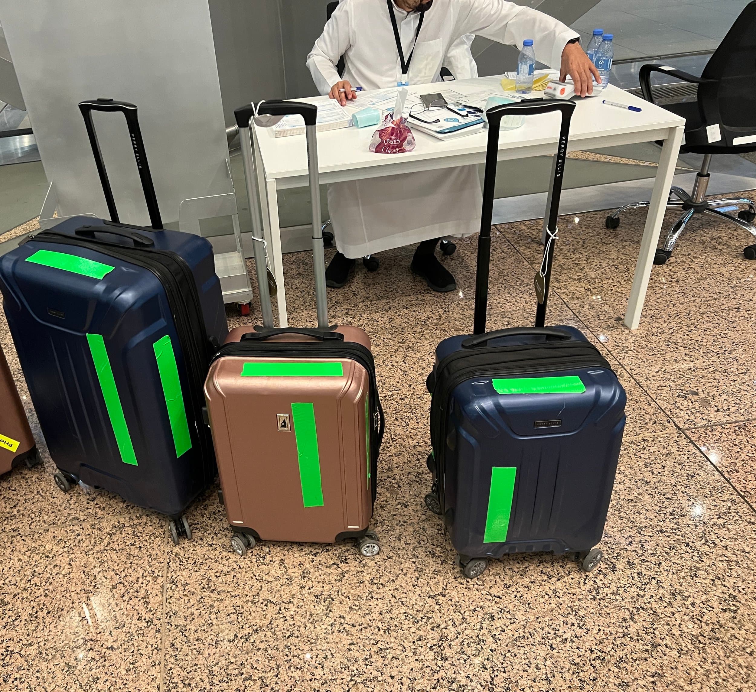 Three suitcases with green luggage tags arranged in front of a person sitting at a desk in an airport.